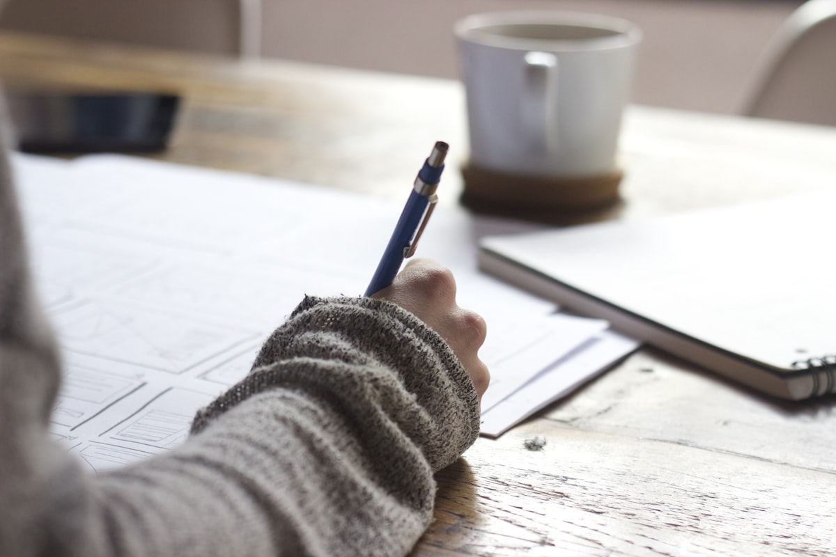 A learner writing notes by hand at a desk, beside a notebook and a mug